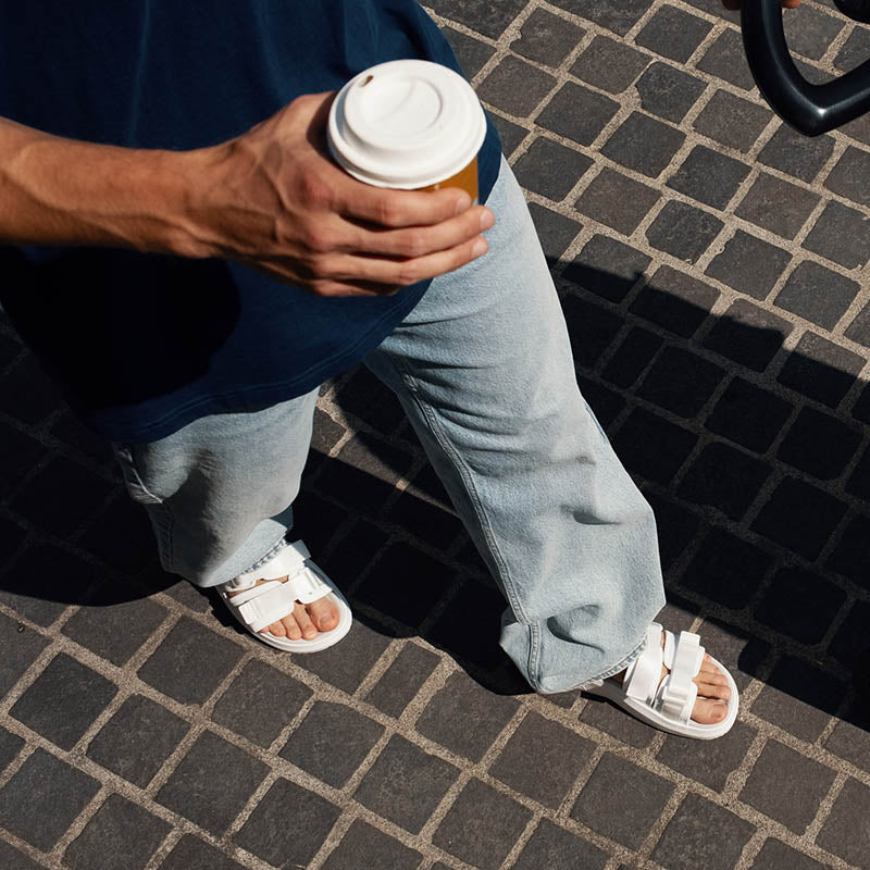 Hvid sporty sandal på en herrefod, med en kaffekop i hånden på et hårdt underlag.